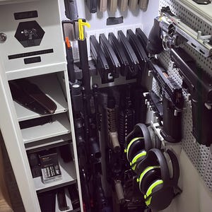 May include: A grey metal gun safe, door ajar, displaying firearms, magazines, and accessories. The safe features multiple shelves and compartments, with items neatly arranged. Includes a pair of black and yellow earmuffs.