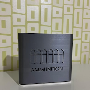 May include: A gray rectangular container with the word "AMMUNITION" in capital letters. Above the text are six bullet illustrations. The container has a white base and is sitting on a white surface.