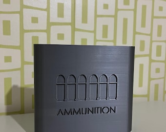 Może przedstawiać: Szary prostokątny pojemnik z napisem "AMMUNITION" wielkimi literami. Nad tekstem znajduje się sześć ilustracji pocisków. Pojemnik ma białą podstawę i stoi na białej powierzchni.