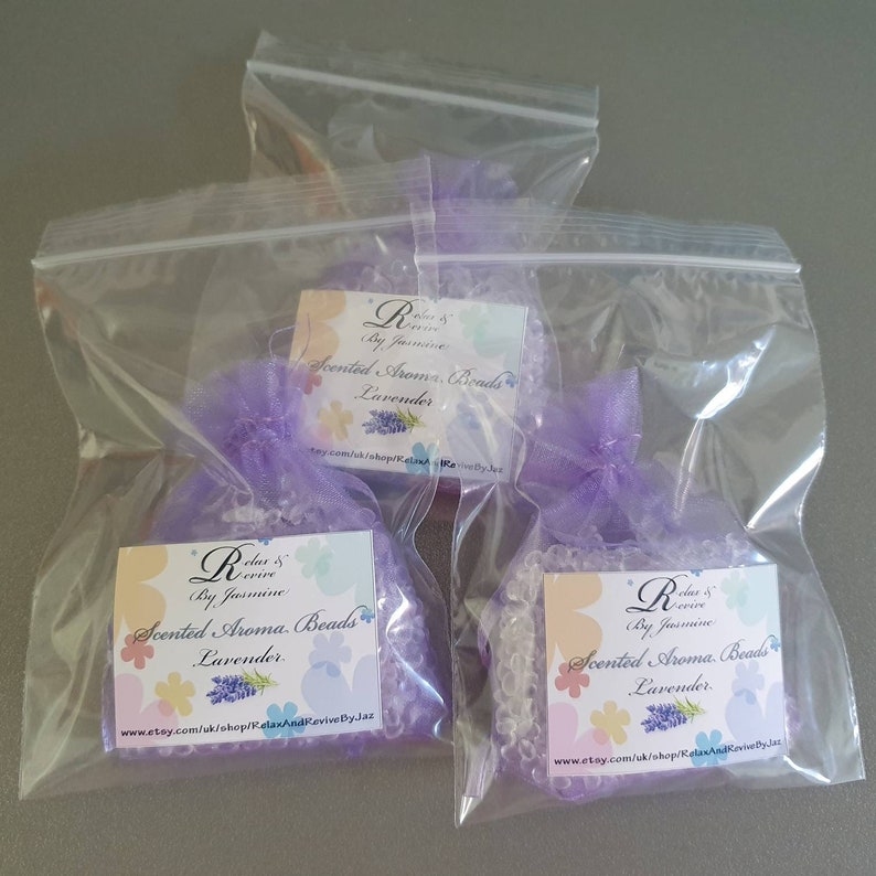 May include: Three clear plastic bags with purple ribbon bows contain lavender scented aroma beads. Each bag has a white label with the text "Relax & Revive by Jasmine Scented Aroma Beads Lavender" and a website address.