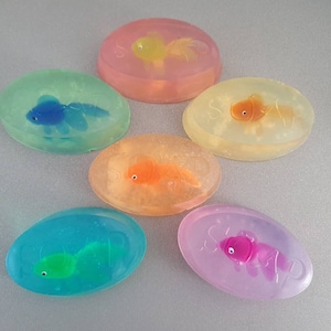 May include: Six oval-shaped soaps with different coloured translucent bases and a small plastic fish figurine inside each. The fish are orange, blue, green, pink, and yellow.