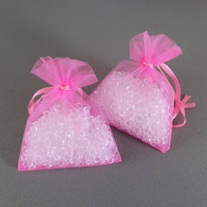 May include: Two pink tulle bags filled with clear, round beads. The bags are tied with a pink ribbon.