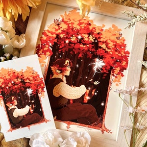 May include: Two framed prints of a fantasy illustration featuring a person with a furry ear and a white sweater sitting under a tree with glowing lights. The tree has red, orange, and yellow leaves. The print on the left is smaller than the print on the right.