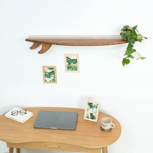 Op de afbeelding: Een houten plank in de vorm van een surfplank met vinnen, gemonteerd aan een witte muur. Daaronder een houten bureau met een laptop, bril, boek en ingelijste kunst. Een mok en een stoel maken de werkruimte compleet. Een plant staat op de plank.