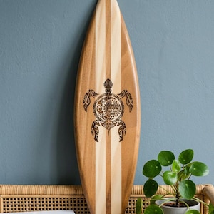 Op de afbeelding: Houten surfboard met een lichtbruin en crèmekleurig gestreept ontwerp. Een gedetailleerd schildpadontwerp is in het midden geëtst. Het surfboard leunt tegen een blauwe muur. Een kleine potplant en boeken staan op de voorgrond.
