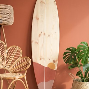 Peut inclure: Planche de surf en bois avec un motif de coucher de soleil peint en nuances de rouge, orange et blanc. La planche de surf est appuyée contre un mur de couleur terre cuite. La planche a un fini grain de bois naturel.