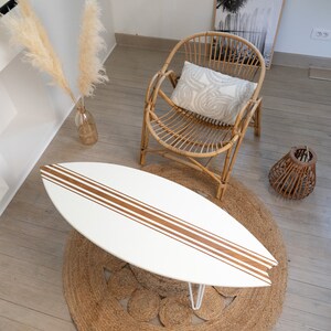 May include: White surfboard coffee table with brown stripes on a jute rug. A wicker chair with a white cushion is in the background.