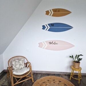 May include: Three wooden surfboard-shaped wall decorations with names written on them. The top surfboard is brown with the name "Hélie" written on it. The middle surfboard is blue with the name "Hugo" written on it. The bottom surfboard is pink with the name "Malice" written on it.