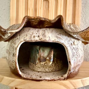 Puede incluir: Un recipiente de cerámica hecho a mano con una forma orgánica única. El exterior presenta un esmalte moteado marrón y crema, con un borde marrón más oscuro que se ensancha hacia afuera. El interior revela una pieza de cerámica más pequeña y texturizada con tonos verdes y marrones. Sobre un soporte de madera.
