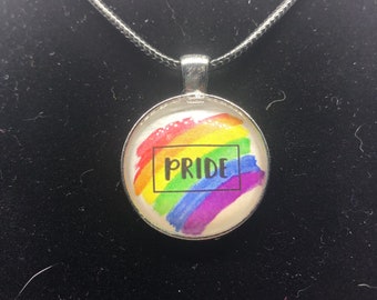 Puede incluir: Un collar de plata con un colgante redondo que presenta un diseño de arcoíris pintado y la palabra "PRIDE" en letras negras.
