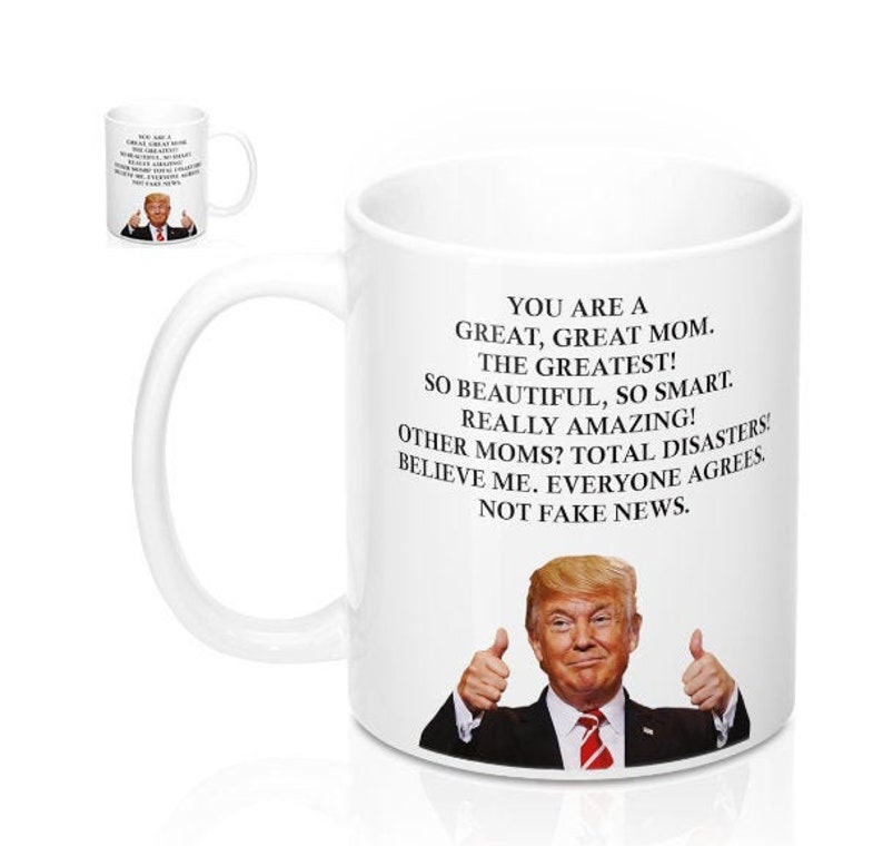 May include: White ceramic coffee mug with a black handle. The mug features a graphic of a man with a thumbs up gesture and the text "YOU ARE A GREAT, GREAT MOM. THE GREATEST! SO BEAUTIFUL, SO SMART. REALLY AMAZING! OTHER MOMS? TOTAL DISASTERS! BELIEVE ME. EVERYONE AGREES. NOT FAKE NEWS."