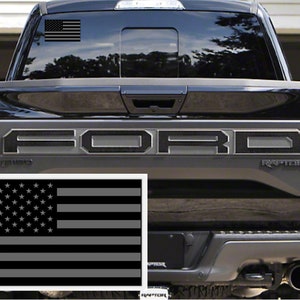 May include: A black Ford Raptor pickup truck with a gray American flag decal on the rear window and a larger gray American flag decal on the tailgate. The truck has a black carbon fiber-style "FORD" lettering on the tailgate.