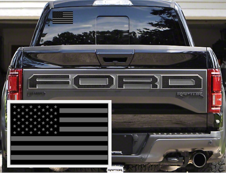 May include: A black Ford Raptor pickup truck with a gray American flag decal on the rear window and a larger gray American flag decal on the tailgate. The truck has a black carbon fiber-style "FORD" lettering on the tailgate.