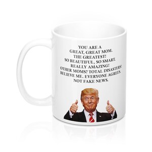 May include: White ceramic coffee mug featuring a black and white image of a man giving a thumbs up. The text on the mug reads: "You are a great, great mom. The greatest! So beautiful, so smart. Really amazing! Other moms? Total disasters! Believe me. Everyone agrees. Not fake news."