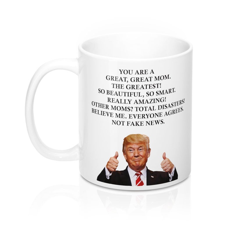May include: White ceramic coffee mug featuring a black and white image of a man giving a thumbs up. The text on the mug reads: "You are a great, great mom. The greatest! So beautiful, so smart. Really amazing! Other moms? Total disasters! Believe me. Everyone agrees. Not fake news."