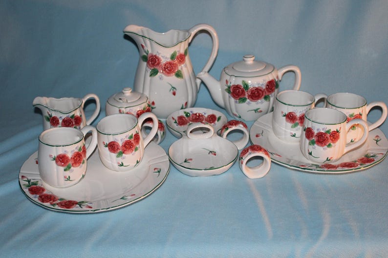 May include: A white ceramic tea set with red rose floral designs. The set includes a teapot, creamer, sugar bowl, cups, saucers, and a pitcher. The set also includes matching napkin rings and a small dish.