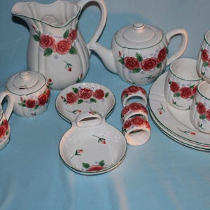 May include: A white ceramic tea set with a floral rose design. The set includes a teapot, creamer, sugar bowl, cups, saucers, and a pitcher. The roses are red with green leaves, and the set has a green trim.