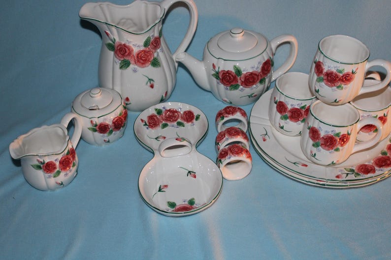 May include: A white ceramic tea set with a floral rose design. The set includes a teapot, creamer, sugar bowl, cups, saucers, and a pitcher. The roses are red with green leaves, and the set has a green trim.