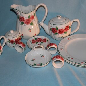 May include: A white ceramic tea set with a floral rose design. The set includes a teapot, creamer, sugar bowl, pitcher, plates, and napkin rings. The roses are red with green leaves and accents. The rims are trimmed with green.