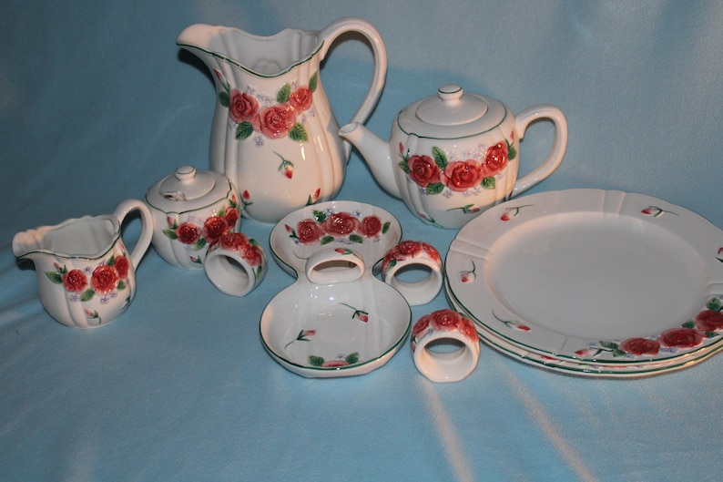 May include: A white ceramic tea set with a floral rose design. The set includes a teapot, creamer, sugar bowl, pitcher, plates, and napkin rings. The roses are red with green leaves and accents. The rims are trimmed with green.