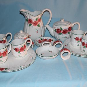 May include: A white ceramic tea set with a floral rose pattern. The set includes a teapot, creamer, sugar bowl, pitcher, cups, saucers, and napkin rings. The roses are red with green leaves and accents.
