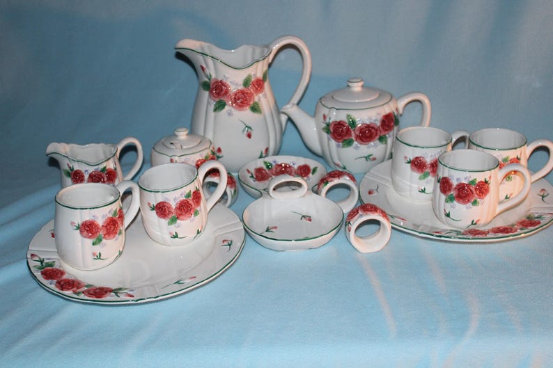 May include: A white ceramic tea set with a floral rose pattern. The set includes a teapot, creamer, sugar bowl, pitcher, cups, saucers, and napkin rings. The roses are red with green leaves and accents.