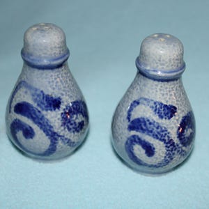 May include: A pair of ceramic salt and pepper shakers. The shakers are light blue with a speckled texture and dark blue, swirled designs. Each shaker has a rounded top with a ring of dark blue and a series of small holes.