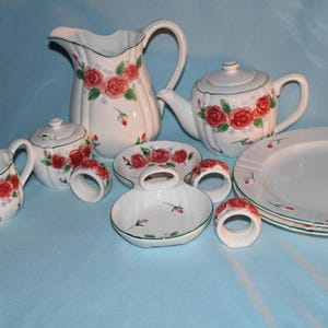 May include: A white ceramic tea set with a floral rose design. The set includes a teapot, creamer, sugar bowl, pitcher, plates, and napkin rings. The roses are red with green leaves and accents. The set is on a light blue surface.