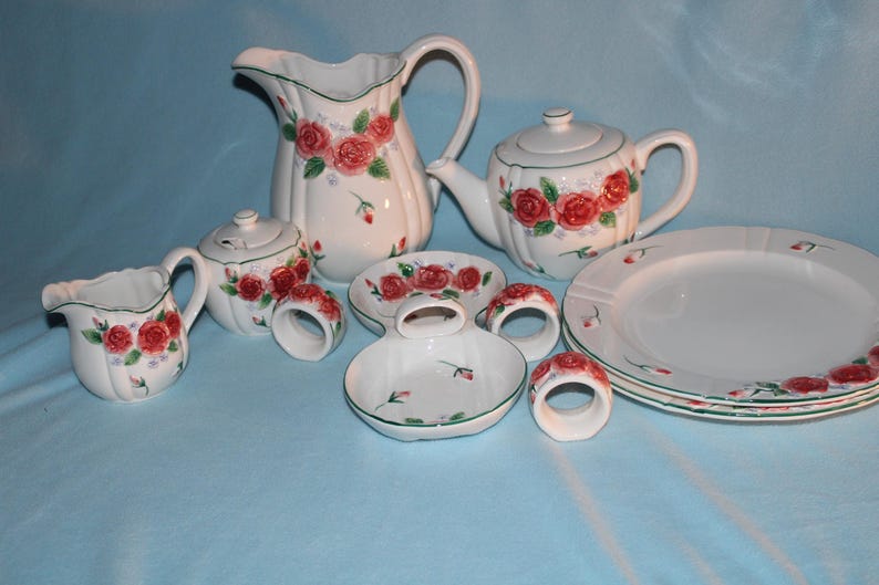 May include: A white ceramic tea set with a floral rose design. The set includes a teapot, creamer, sugar bowl, pitcher, plates, and napkin rings. The roses are red with green leaves and accents. The set is on a light blue surface.