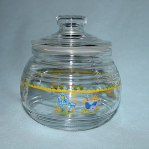 May include: A clear glass jar with a lid, decorated with a yellow, green, and blue floral design. The jar has a round shape and a wide mouth.