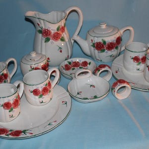 May include: A white ceramic tea set with a floral rose design. The set includes a pitcher, teapot, sugar bowl, creamer, cups, saucers, ashtrays, and napkin rings. The roses are red with green leaves and stems.