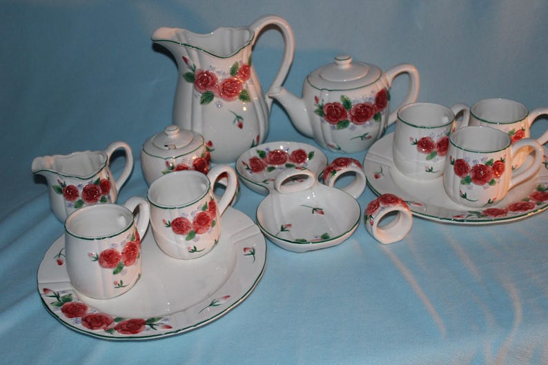 May include: A white ceramic tea set with a floral rose design. The set includes a pitcher, teapot, sugar bowl, creamer, cups, saucers, ashtrays, and napkin rings. The roses are red with green leaves and stems.