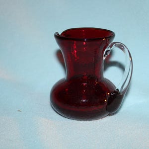 May include: A small, ruby-red glass pitcher with a clear handle. The pitcher has a bulbous base and a slightly flared rim. The glass has a textured, crackled appearance, and the handle is attached near the top and bottom of the pitcher.