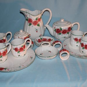 May include: A white ceramic tea set with red rose floral designs. The set includes a teapot, creamer, sugar bowl, cups, saucers, and a pitcher. The set also includes matching napkin rings and a small dish.