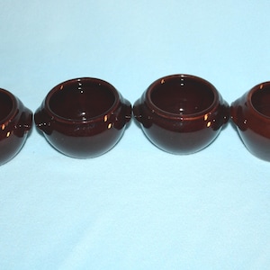 May include: Four small, round, brown ceramic bowls with handles. The bowls are arranged in a row, each with a glossy finish. The interior of each bowl is a darker shade of brown.