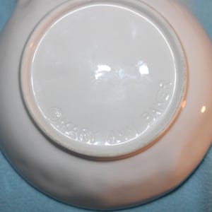 May include: A white ceramic dish with a slightly curved rim. The underside of the dish is visible, featuring a circular indentation and embossed text that reads "MARY ANN BAKER". The dish is set against a light blue background.