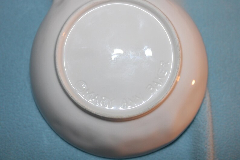 May include: A white ceramic dish with a slightly curved rim. The underside of the dish is visible, featuring a circular indentation and embossed text that reads "MARY ANN BAKER". The dish is set against a light blue background.