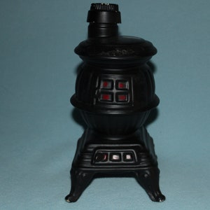 May include: Black ceramic stove-shaped bottle with a red glass window. The bottle has a black plastic lid and sits on a blue surface.