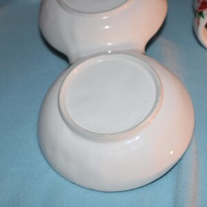 May include: White ceramic serving dish with two connected bowls. The bottom of the dish has a circular base. Other ceramic pieces with floral designs are visible in the background.