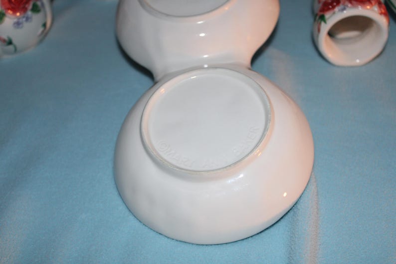 May include: White ceramic serving dish with two connected bowls. The bottom of the dish has a circular base. Other ceramic pieces with floral designs are visible in the background.