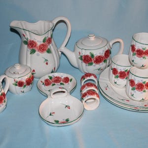 May include: A white ceramic tea set with a red rose pattern. The set includes a teapot, creamer, sugar bowl, pitcher, cups, saucers, and napkin rings. Green trim accents the edges of the pieces.
