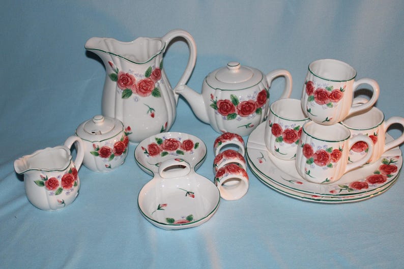 May include: A white ceramic tea set with a red rose pattern. The set includes a teapot, creamer, sugar bowl, pitcher, cups, saucers, and napkin rings. Green trim accents the edges of the pieces.