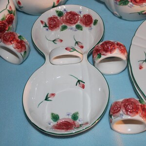 May include: White ceramic dinnerware set with a red rose pattern. Includes plates, serving dishes, a teapot, and napkin rings. The rims are trimmed with green. The set is designed for serving meals.