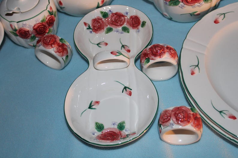 May include: White ceramic dinnerware set with a red rose pattern. Includes plates, serving dishes, a teapot, and napkin rings. The rims are trimmed with green. The set is designed for serving meals.
