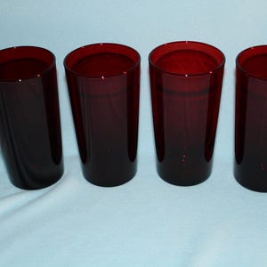 May include: Four tall, ruby-red drinking glasses. The glasses are cylindrical with a slightly flared top. The color is consistent throughout, with a deep, rich hue. The glasses are arranged in a row on a light-colored surface.