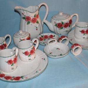 May include: A white ceramic tea set with red rose floral designs. The set includes a teapot, creamer, sugar bowl, cups, saucers, and a pitcher. Green trim accents the edges of the pieces. Napkin rings and a soap dish are also included.