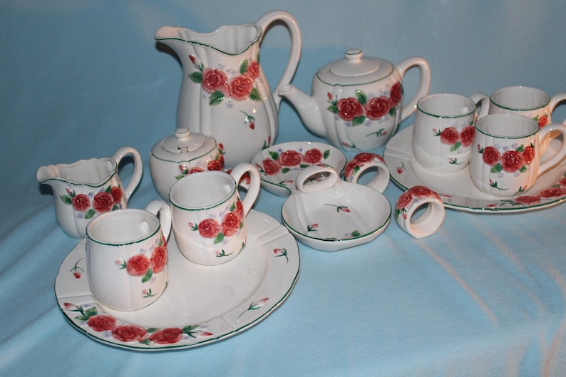 May include: A white ceramic tea set with red rose floral designs. The set includes a teapot, creamer, sugar bowl, cups, saucers, and a pitcher. Green trim accents the edges of the pieces. Napkin rings and a soap dish are also included.