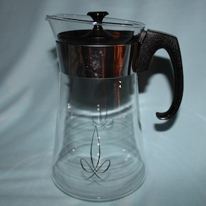 May include: A clear glass coffee carafe with a black handle, lid, and trim. The carafe has a flared shape and a decorative black design on the glass. The carafe is designed to hold hot liquids.