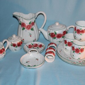 May include: A white ceramic tea set with a floral rose design. The set includes a teapot, creamer, sugar bowl, cups, saucers, plates, and napkin rings. The roses are red with green leaves, and the rims are trimmed in green.