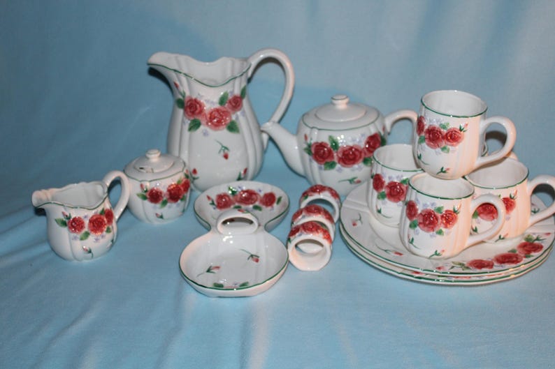 May include: A white ceramic tea set with a floral rose design. The set includes a teapot, creamer, sugar bowl, cups, saucers, plates, and napkin rings. The roses are red with green leaves, and the rims are trimmed in green.